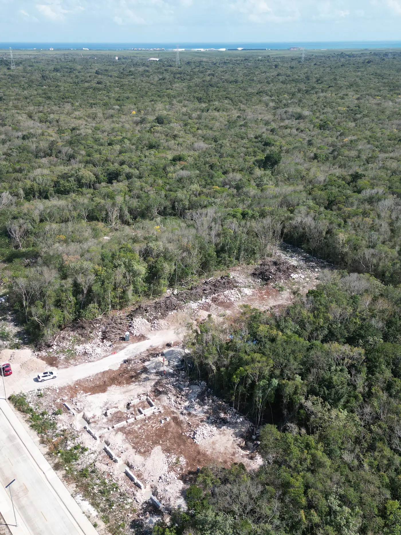 Aerial view of a cleared industrial lot beside a paved road, concrete footings and cut brush surrounded by dense jungle