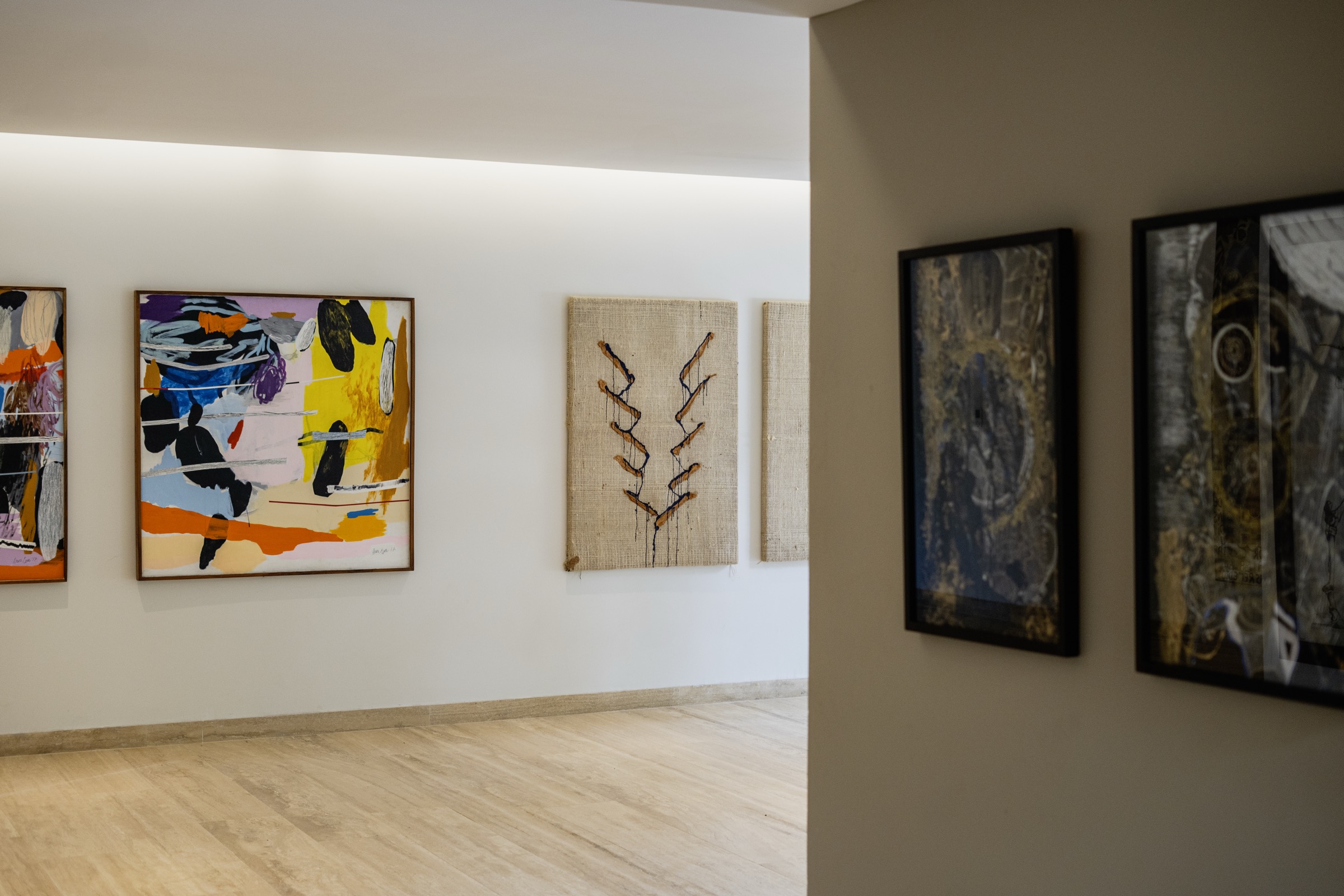 Photograph of a gallery corridor hung with framed abstract canvases and two burlap works on a pale plank floor